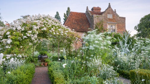 A brick path leads through masses of white flowers and under an arch covered in white roses in the White Garden at Sissinghurst Castle Garden, Kent, in June